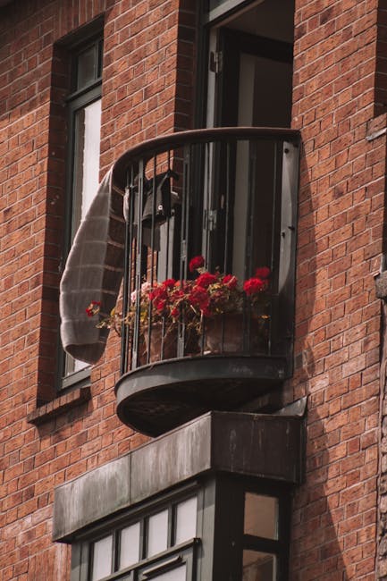 A modern multi-storey building featuring numerous balconies, each decorated with a variety of plants, shrubs, and small trees. The greenery includes a mix of evergreen and flowering species, with some