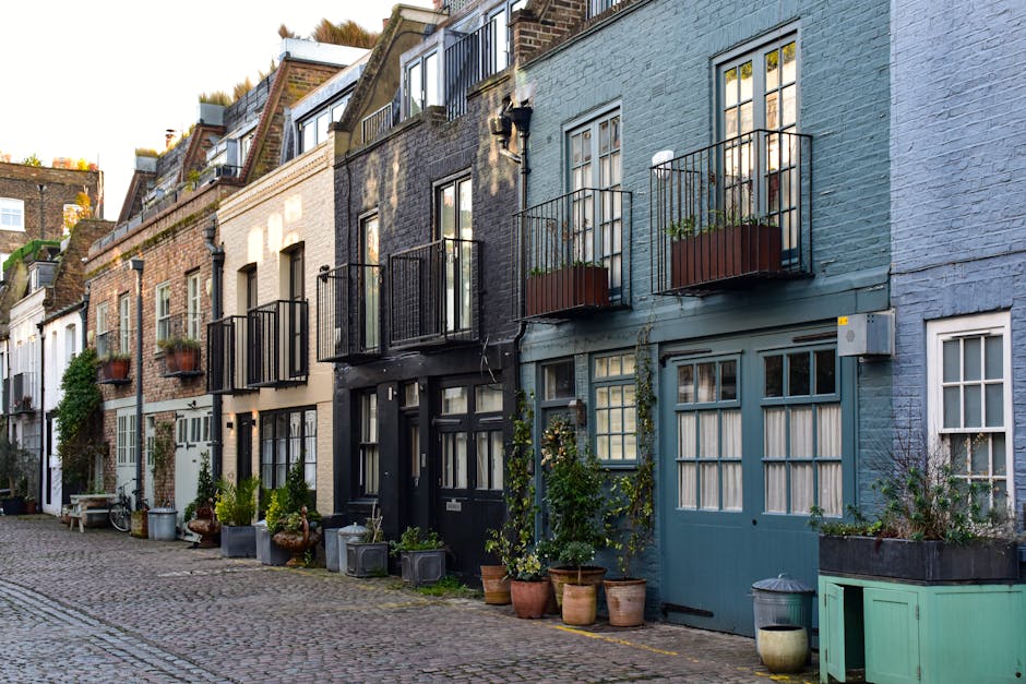 A row of townhouses along a cobblestone street, each featuring small front gardens filled with potted plants, shrubs, and climbing vines. The houses have diverse exterior colors including blue, black,